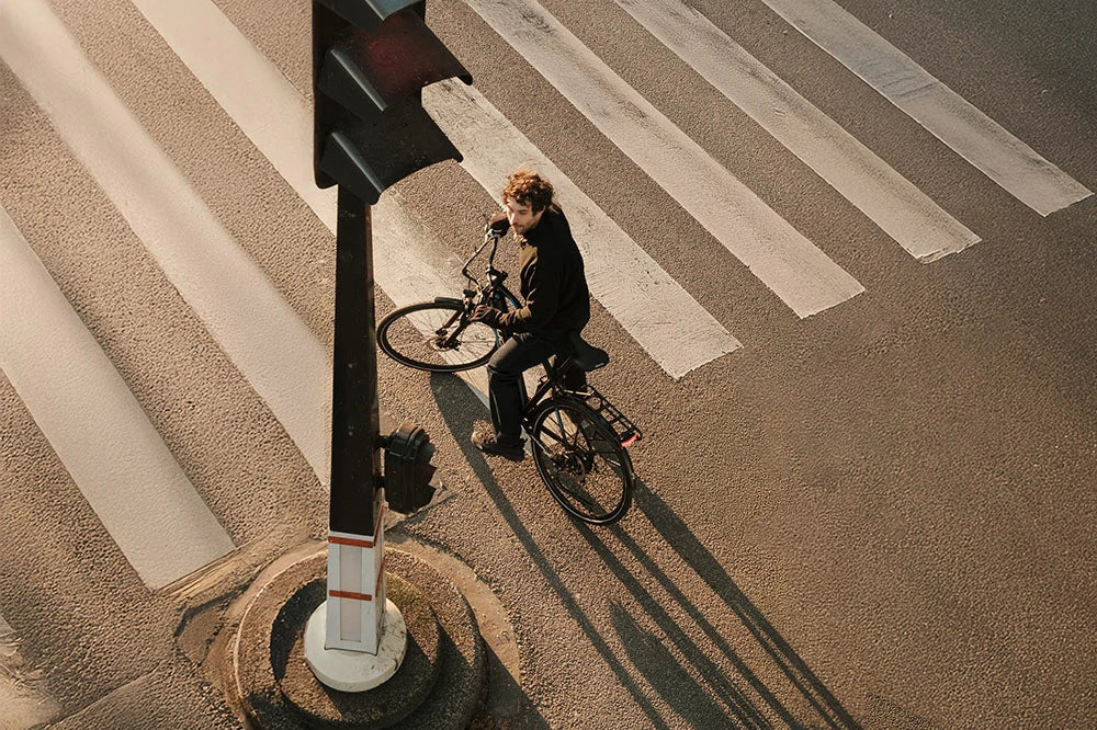 Aerial shot of a cyclist on the DUOTTS C29 Lite electric bike waiting at a city crosswalk during sunset, showcasing its sleek black frame, rear rack, and urban commuter design
